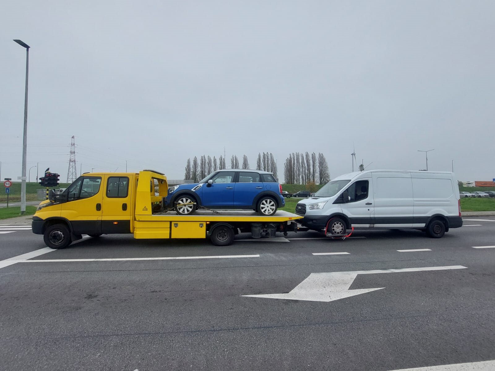 Yellow flatbed recovery truck transporting a blue Mini and towing a white van